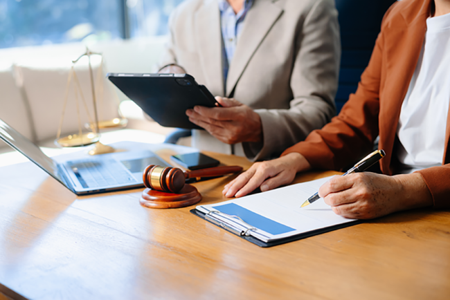 Two people in business clothing sitting at a table with a clipboard, a gavel, a laptop, a tablet, and balance scales.